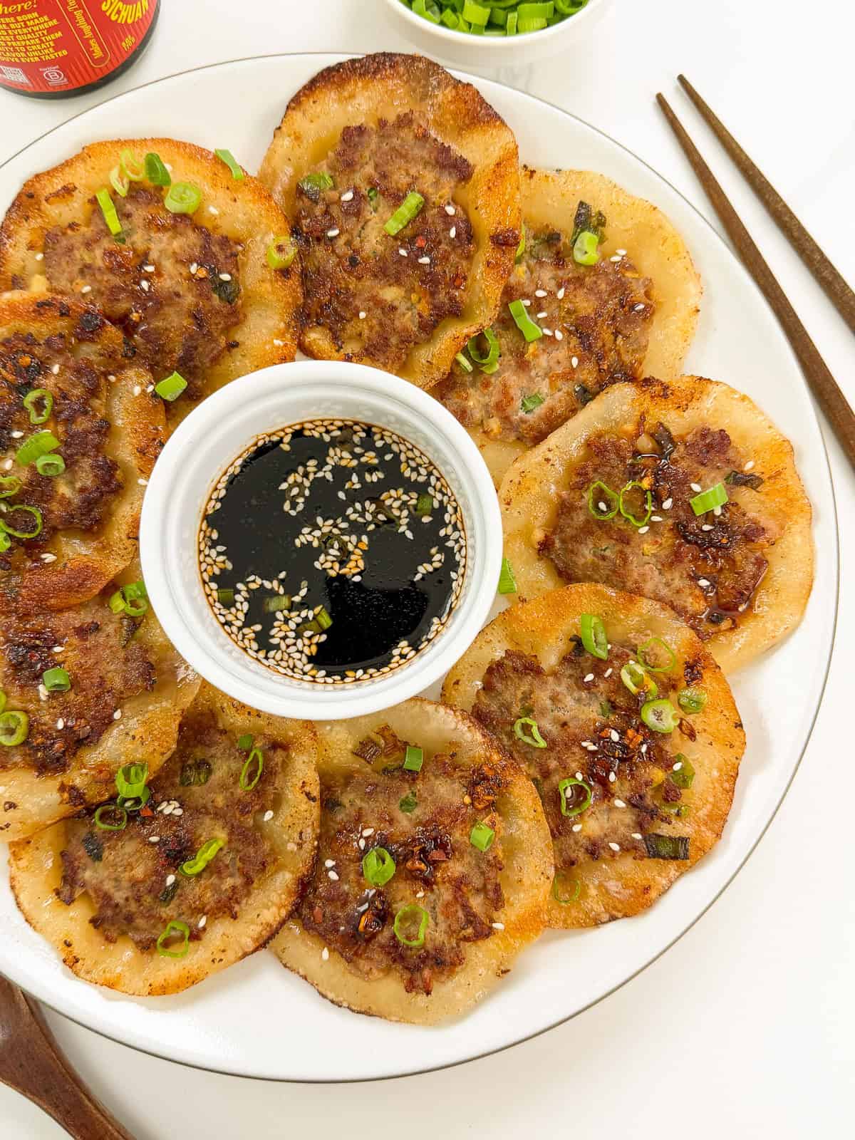 A plate of golden Smashed Lemongrass Pork Dumplings is arranged in a circle around a bowl of soy dipping sauce, garnished with sliced green onions and sesame seeds. Chopsticks are placed beside the plate.