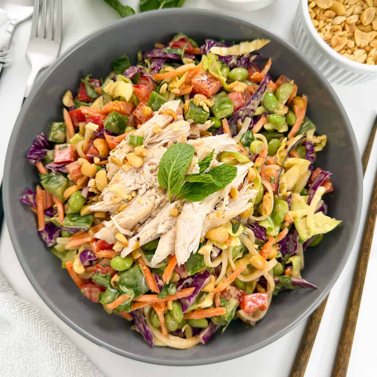 A vibrant Thai Noodle Salad with Peanut Sauce featuring shredded chicken, red cabbage, carrots, bell peppers, edamame, and peanuts, garnished with mint. Chopsticks and a bowl of peanuts sit beside this colorful dish.