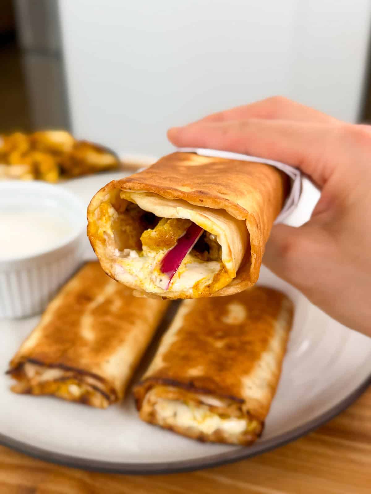 A hand holds a crispy, golden-brown Viral Chicken Shawarma Wrap filled with visible red onions, melted cheese, and other ingredients. Two more wraps and a small bowl of white dipping sauce are on a plate in the background.