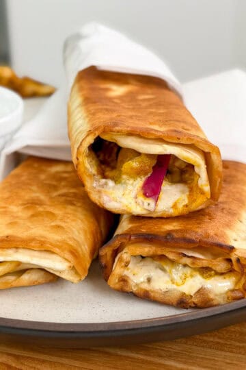 Three crispy, golden-brown Viral Chicken Shawarma Wraps filled with spiced chicken, creamy sauce, and sliced vegetables are stacked on a plate, partially wrapped in white paper, with a small cup of sauce on the side.