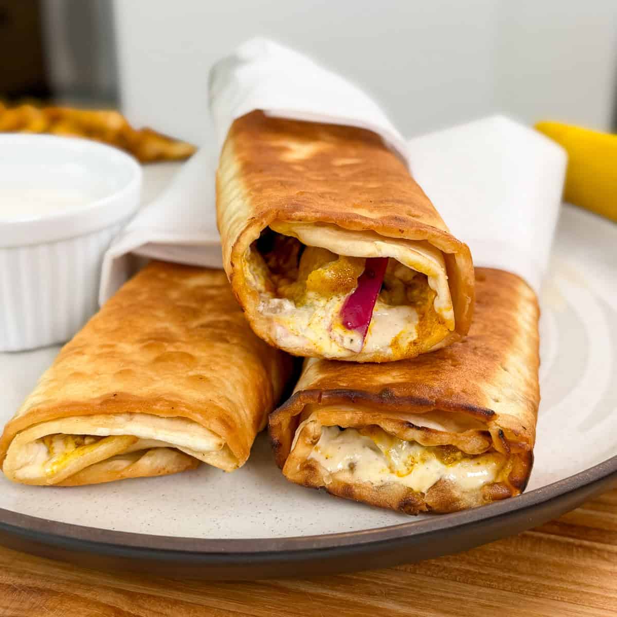 Three crispy, golden-brown Viral Chicken Shawarma Wraps filled with spiced chicken, creamy sauce, and sliced vegetables are stacked on a plate, partially wrapped in white paper, with a small cup of sauce on the side.
