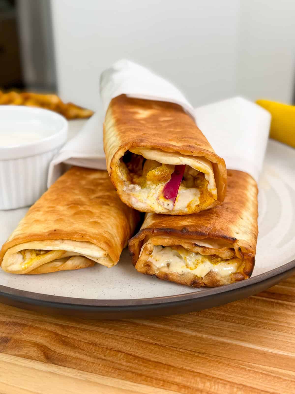 Three crispy, golden-brown Viral Chicken Shawarma Wraps filled with chicken, vegetables, and sauce are stacked on a plate, with a small dish of white dipping sauce in the background.
