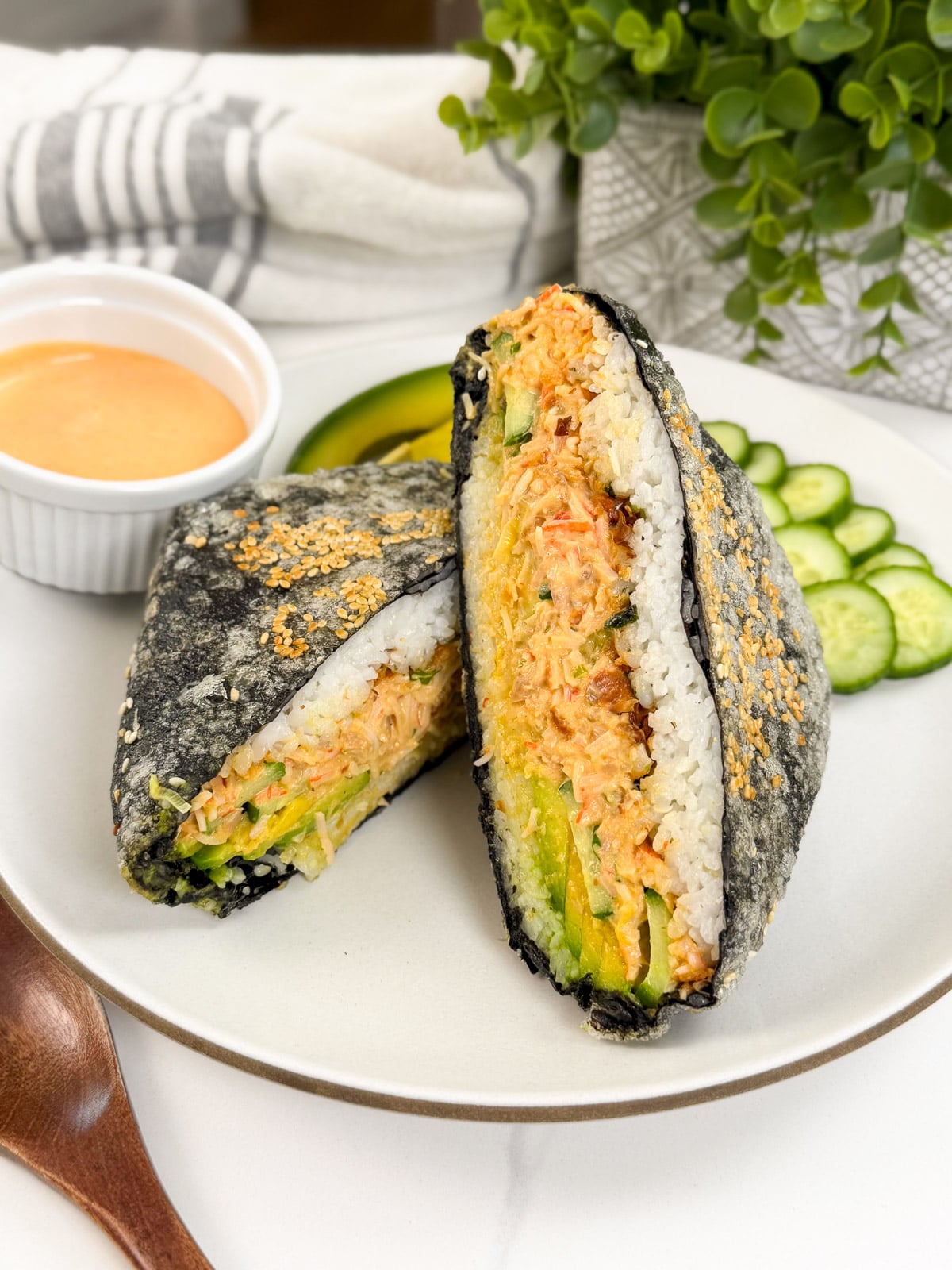 On a plate, a sushi sandwich wrapped in nori and filled with shredded crab, avocado, and cucumber, reminiscent of a California Roll, is cut in half. Sliced cucumber, dipping sauce, and plant decor appear in the background.