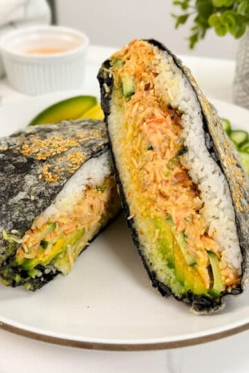 A California Roll-inspired sushi sandwich filled with shredded crab, avocado, and cucumber is wrapped in seaweed and white rice, cut in half, and served on a white plate with cucumber slices and a small dipping sauce in the background.