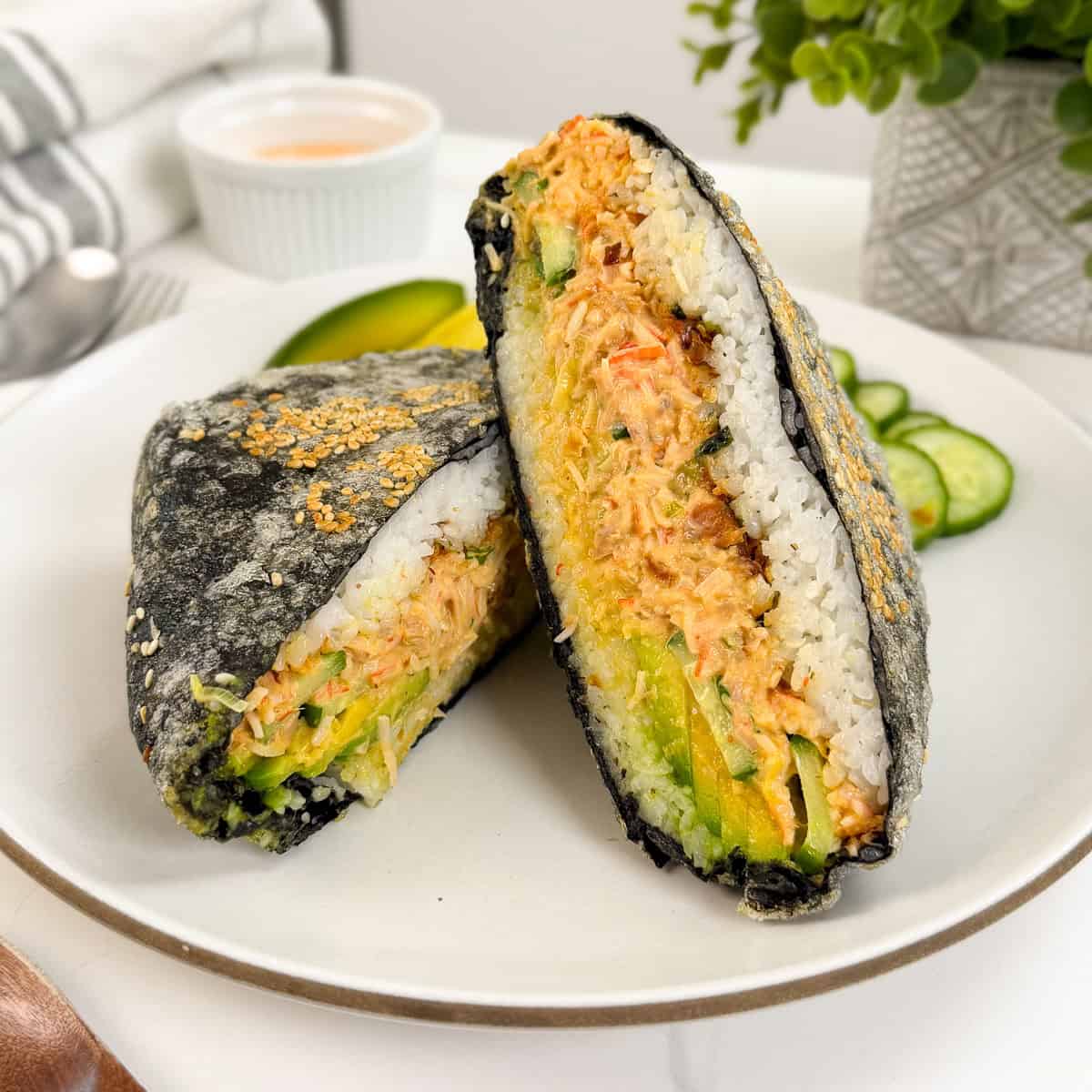 A California Roll-inspired sushi sandwich filled with shredded crab, avocado, and cucumber is wrapped in seaweed and white rice, cut in half, and served on a white plate with cucumber slices and a small dipping sauce in the background.