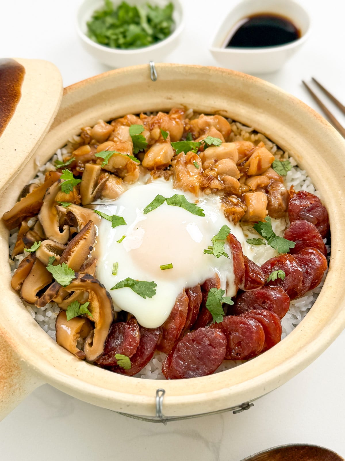 A Cantonese Claypot Rice dish featuring rice in a clay pot, topped with sliced Chinese sausage, mushrooms, diced chicken, and a soft-cooked egg. Chopped herbs garnish the dish, with bowls of sauce and herbs plus chopsticks on the side.