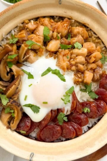A Cantonese Claypot Rice dish filled with rice, topped with sliced Chinese sausage, mushrooms, diced chicken in sauce, a soft-cooked egg, and fresh cilantro. Chopsticks are placed beside the pot for an authentic touch.