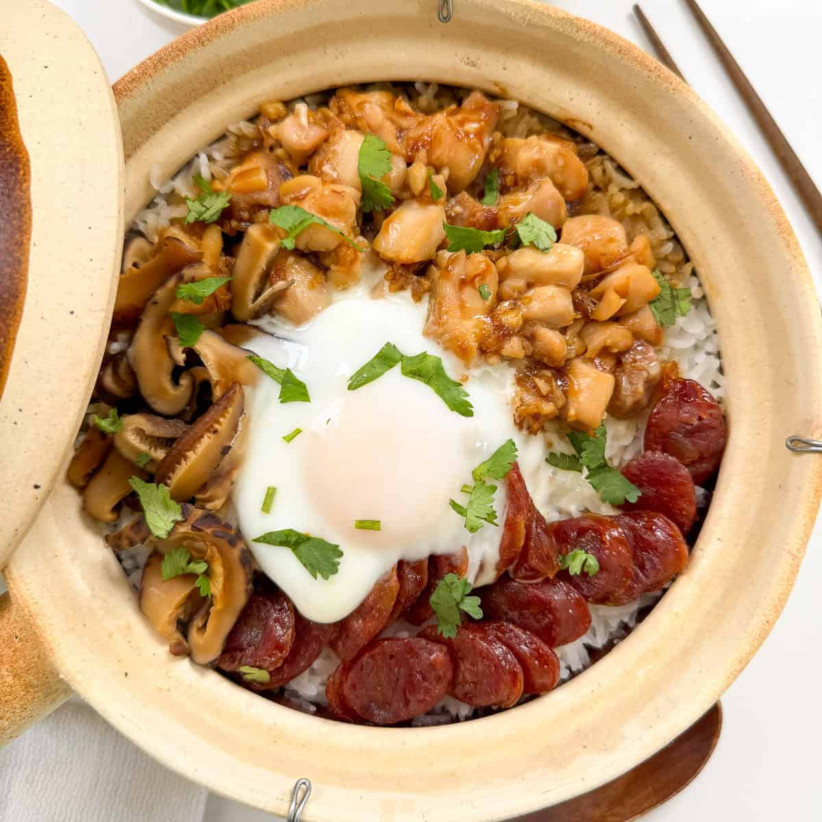 A Cantonese Claypot Rice dish filled with rice, topped with sliced Chinese sausage, mushrooms, diced chicken in sauce, a soft-cooked egg, and fresh cilantro. Chopsticks are placed beside the pot for an authentic touch.