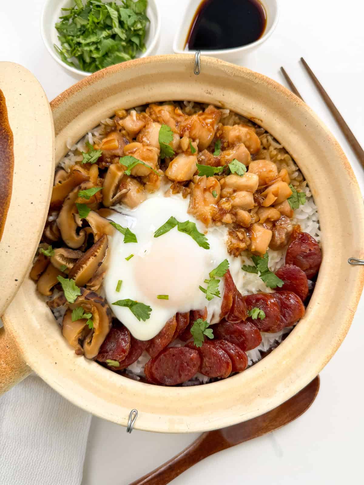 A Cantonese Claypot Rice dish filled with rice, topped with sliced Chinese sausage, chicken, mushrooms, and a poached egg, garnished with chopped cilantro. Small bowls of soy sauce and cilantro sit nearby, alongside chopsticks and a spoon.