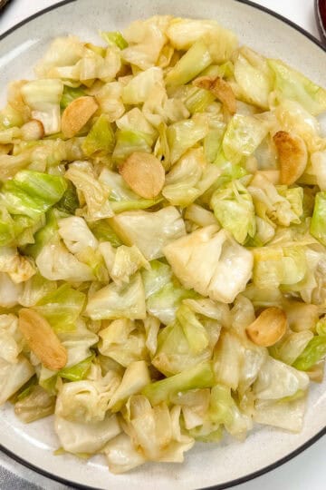 A plate of Chinese Cabbage Stir-Fry with Garlic, featuring tender cabbage and sliced garlic, is served on a white dish with a dark rim. Chopsticks and a wooden spoon are placed beside the plate.