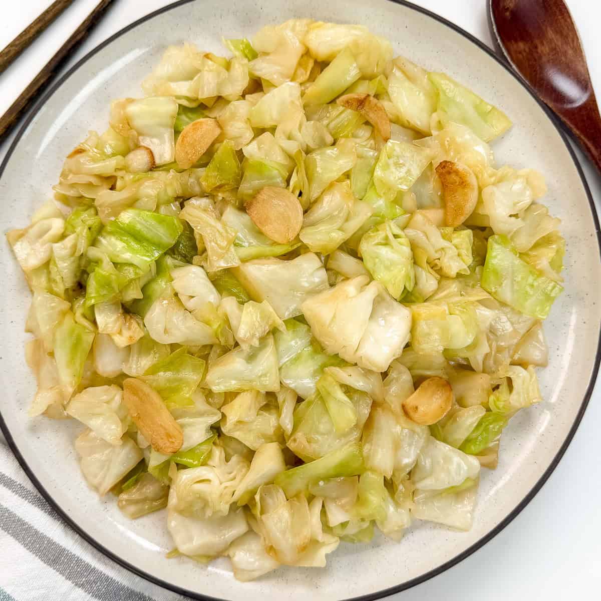 A plate of Chinese Cabbage Stir-Fry with Garlic, featuring tender cabbage and sliced garlic, is served on a white dish with a dark rim. Chopsticks and a wooden spoon are placed beside the plate.