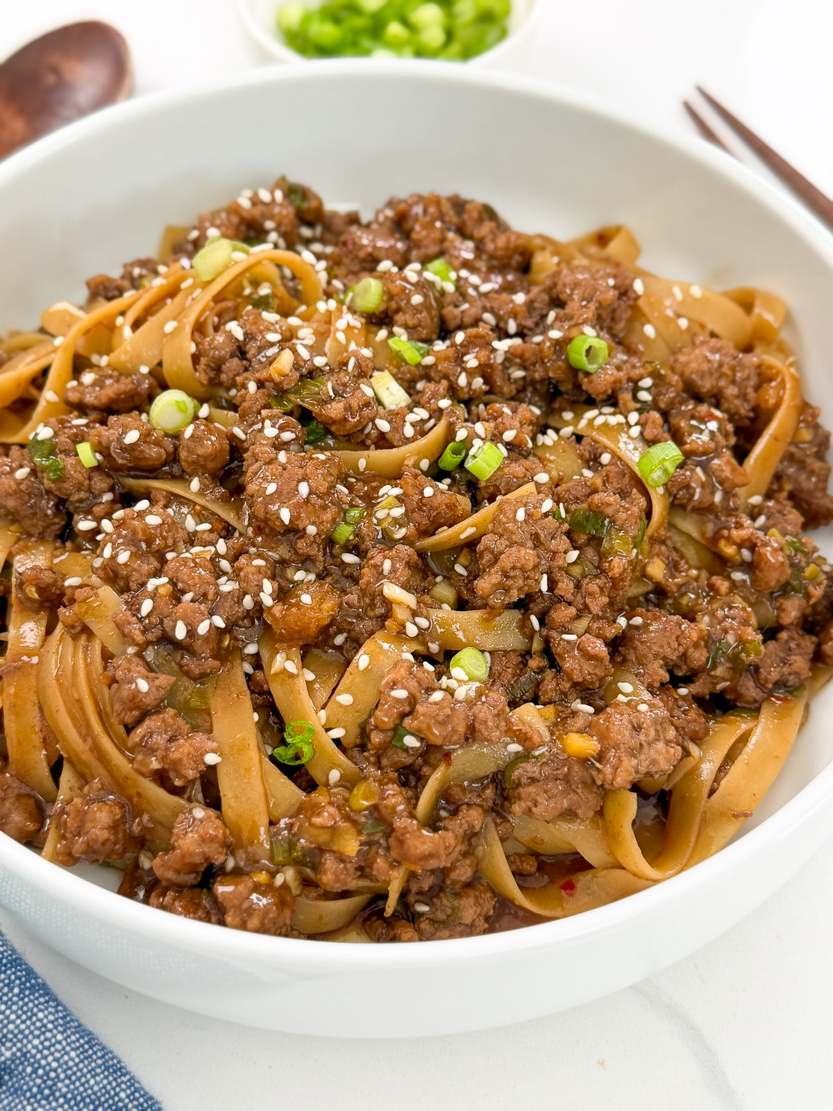 A white bowl filled with Mongolian Ground Beef Noodles, topped with seasoned ground beef, chopped green onions, and sprinkled with white sesame seeds. A small bowl of chopped green onions is in the background.