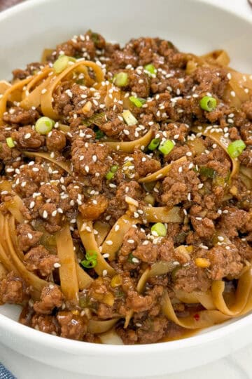 A white bowl filled with Mongolian Ground Beef Noodles, topped with chopped green onions and sprinkled with sesame seeds. A fork is partially visible beside the bowl.