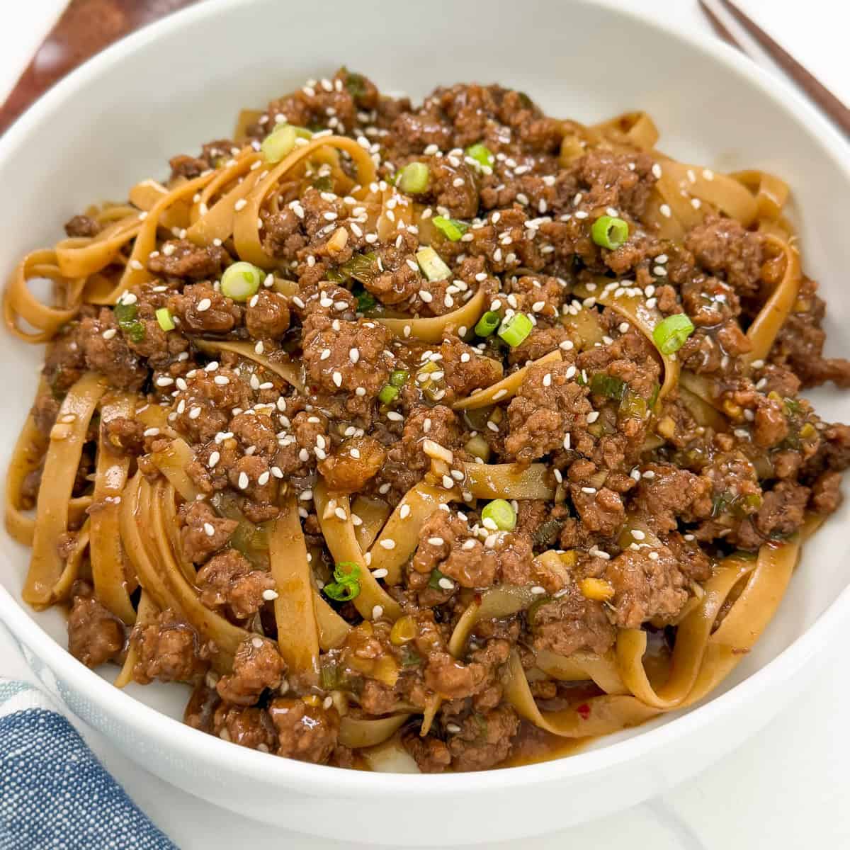 A white bowl filled with Mongolian Ground Beef Noodles, topped with chopped green onions and sprinkled with sesame seeds. A fork is partially visible beside the bowl.
