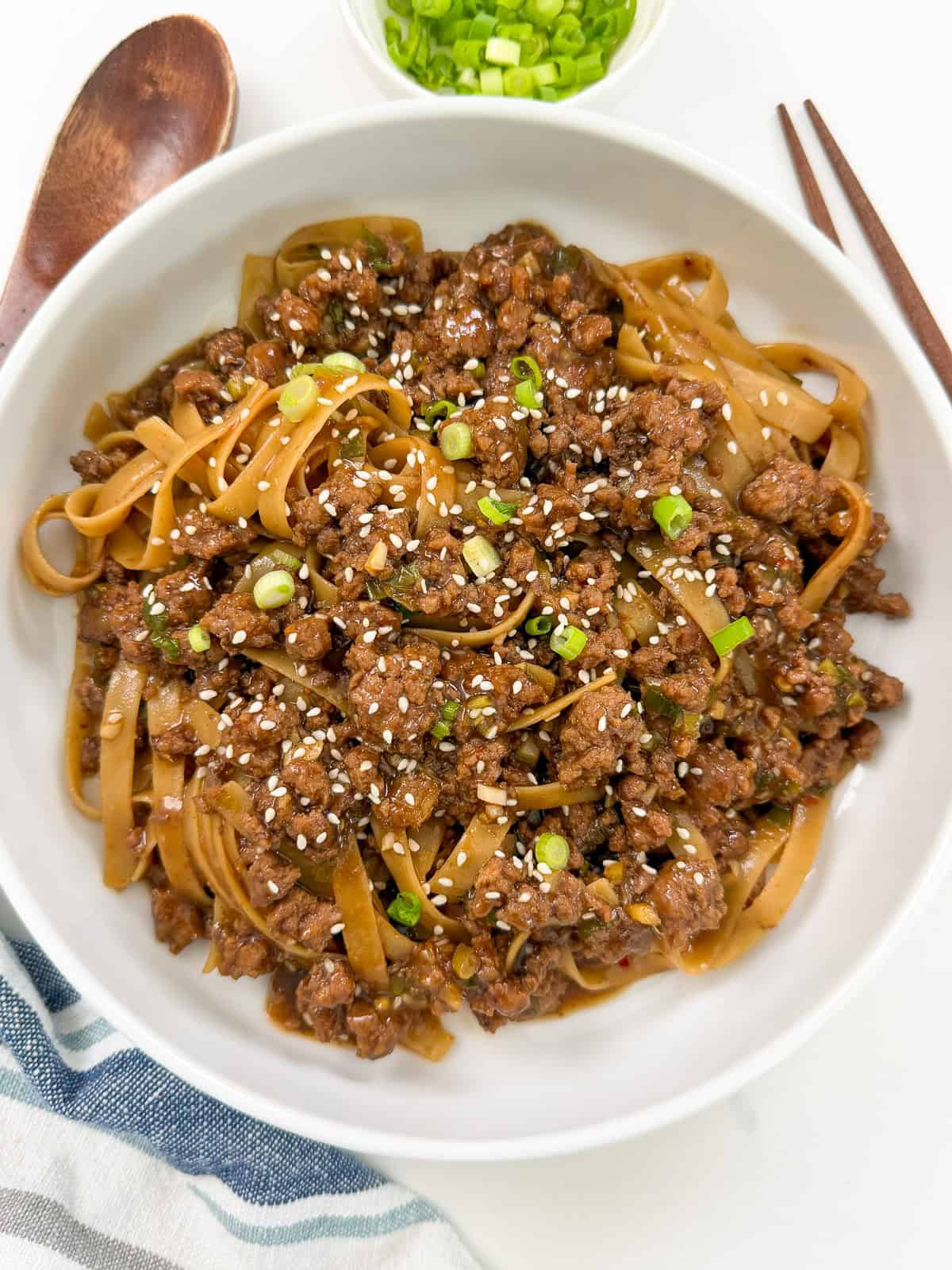 A white bowl filled with saucy Mongolian Ground Beef Noodles and rice noodles, garnished with chopped green onions and sesame seeds. Wooden chopsticks and a spoon are beside the bowl, with a small dish of green onions in the background.