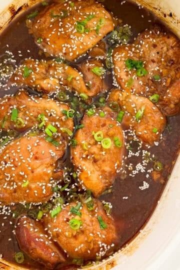Slow Cooker Honey Garlic Chicken thighs cooked in a dark, rich sauce, garnished with chopped green onions and sesame seeds, served in a white slow cooker.