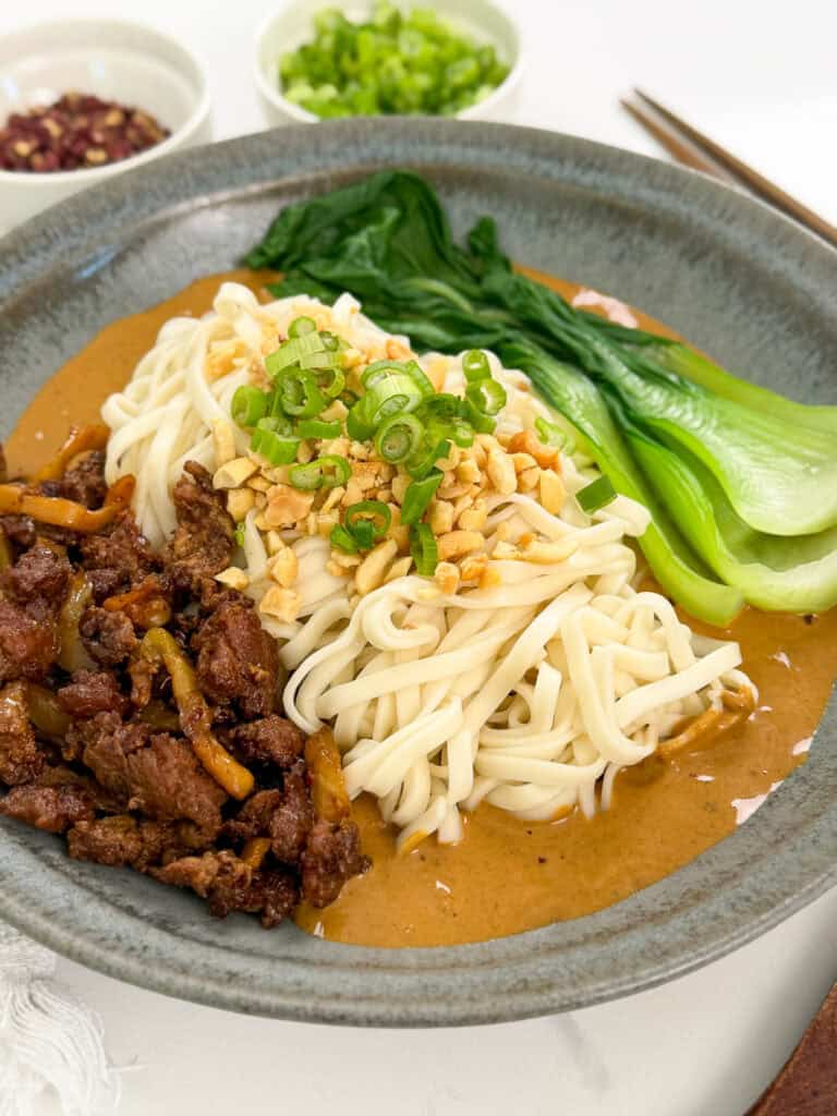 A bowl of Spicy Dan Dan Noodles topped with chopped peanuts, green onions, sliced beef, and bok choy, served with a creamy sauce. Small bowls of chopped green onions and other toppings are in the background.