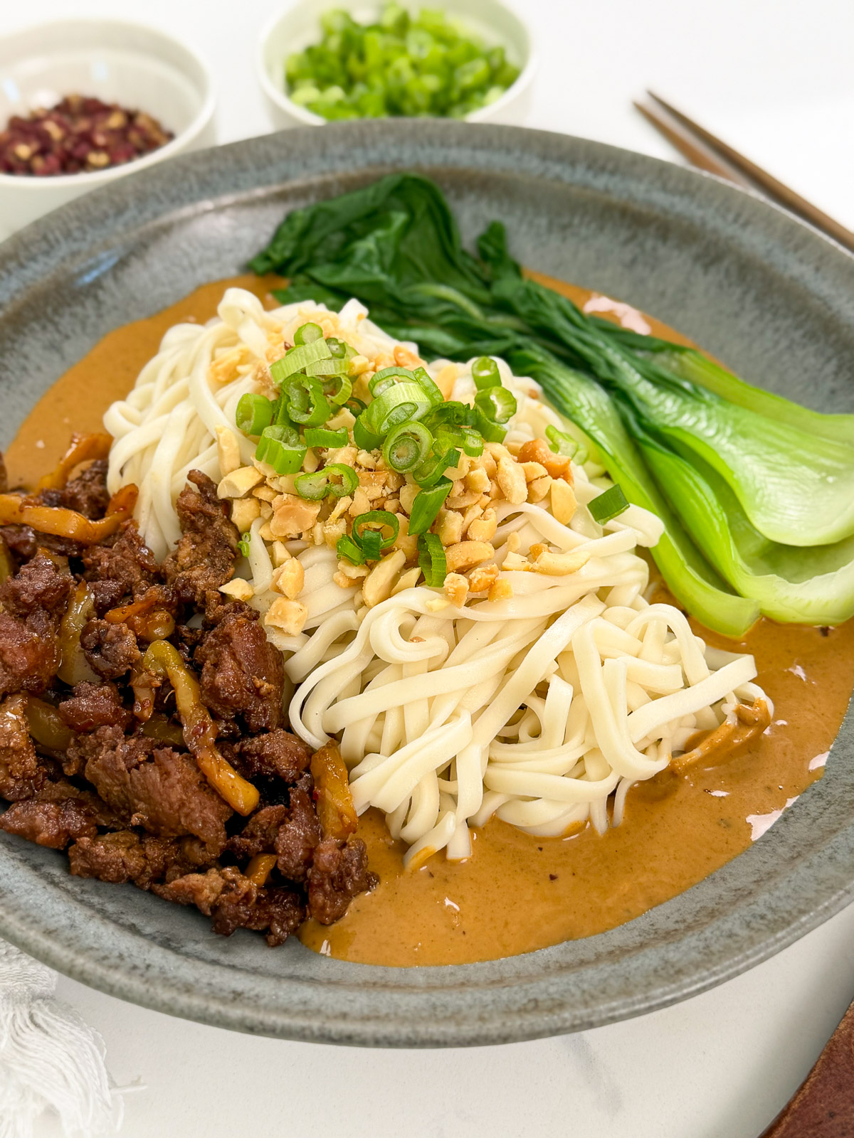 A bowl of Spicy Dan Dan Noodles topped with chopped peanuts, green onions, sliced beef, and bok choy, served with a creamy sauce. Small bowls of chopped green onions and other toppings are in the background.