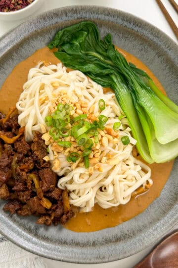 A bowl of Spicy Dan Dan Noodles topped with minced meat, baby bok choy, chopped peanuts, green onions, and sauce, served with chopsticks and a wooden spoon on the side.