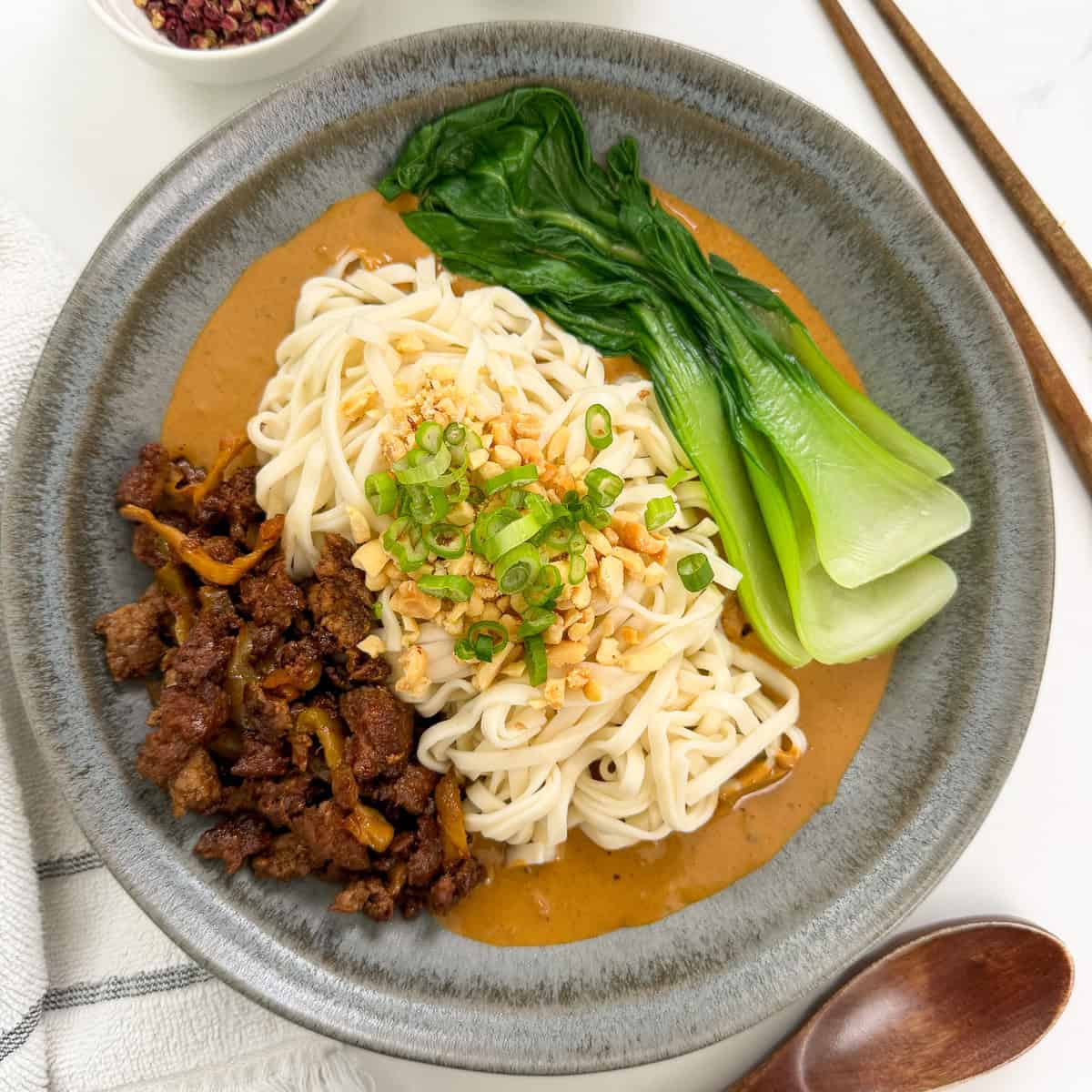 A bowl of Spicy Dan Dan Noodles topped with minced meat, baby bok choy, chopped peanuts, green onions, and sauce, served with chopsticks and a wooden spoon on the side.