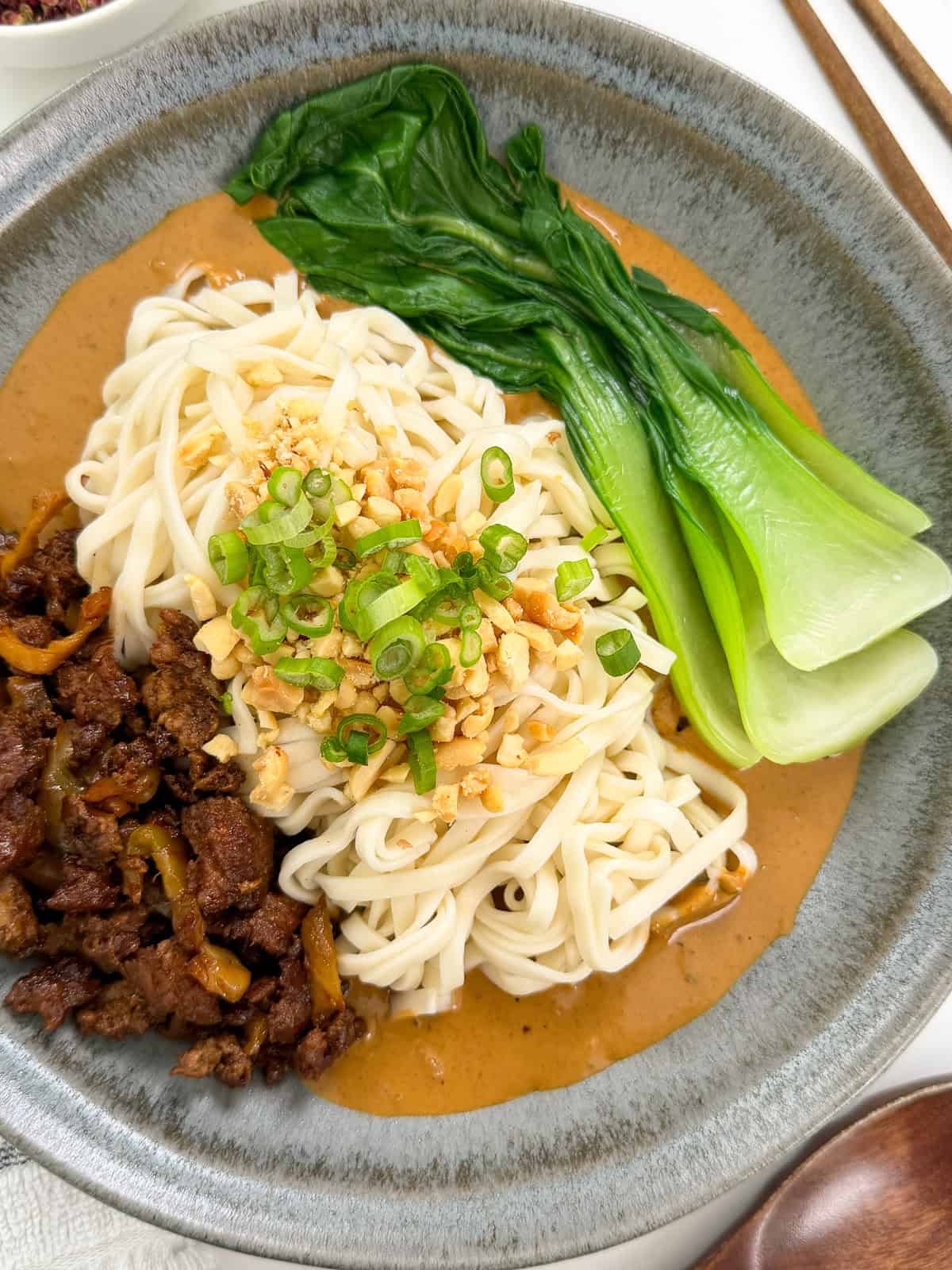A bowl of Spicy Dan Dan Noodles with ground meat, bok choy, creamy sauce, chopped peanuts, and sliced green onions, served in a gray dish with chopsticks nearby.
