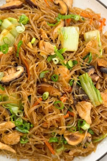 A plate of Stir-Fry Glass Vermicelli Noodles with sliced mushrooms, chicken pieces, bok choy, shredded carrots, and chopped green onions, served on a white dish with chopsticks on the side.