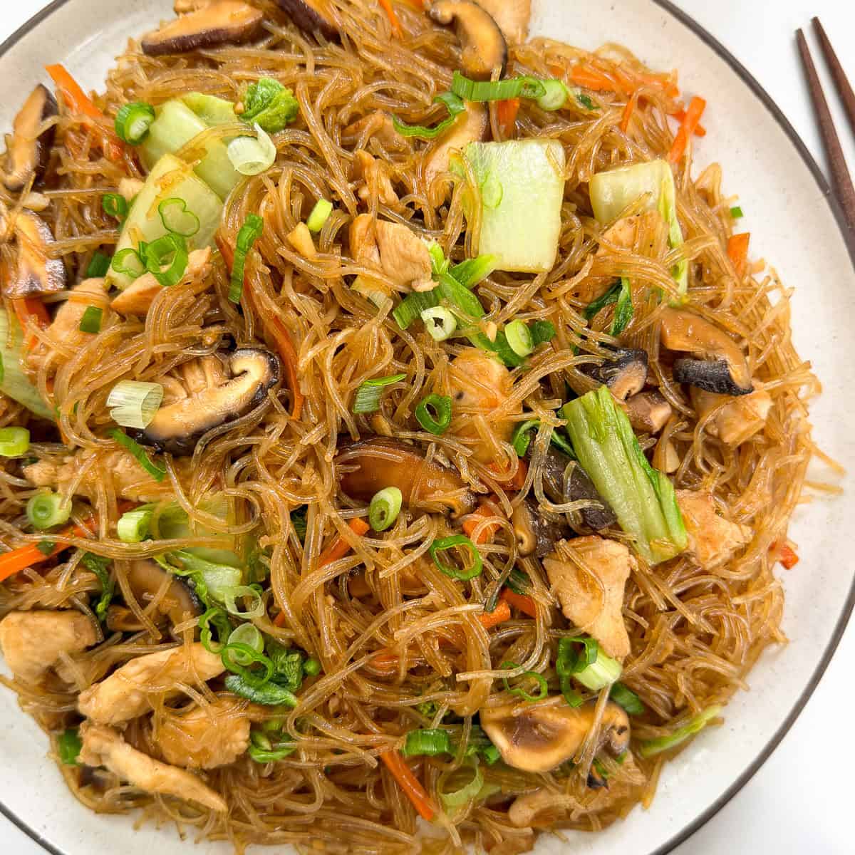 A plate of Stir-Fry Glass Vermicelli Noodles with sliced mushrooms, chicken pieces, bok choy, shredded carrots, and chopped green onions, served on a white dish with chopsticks on the side.