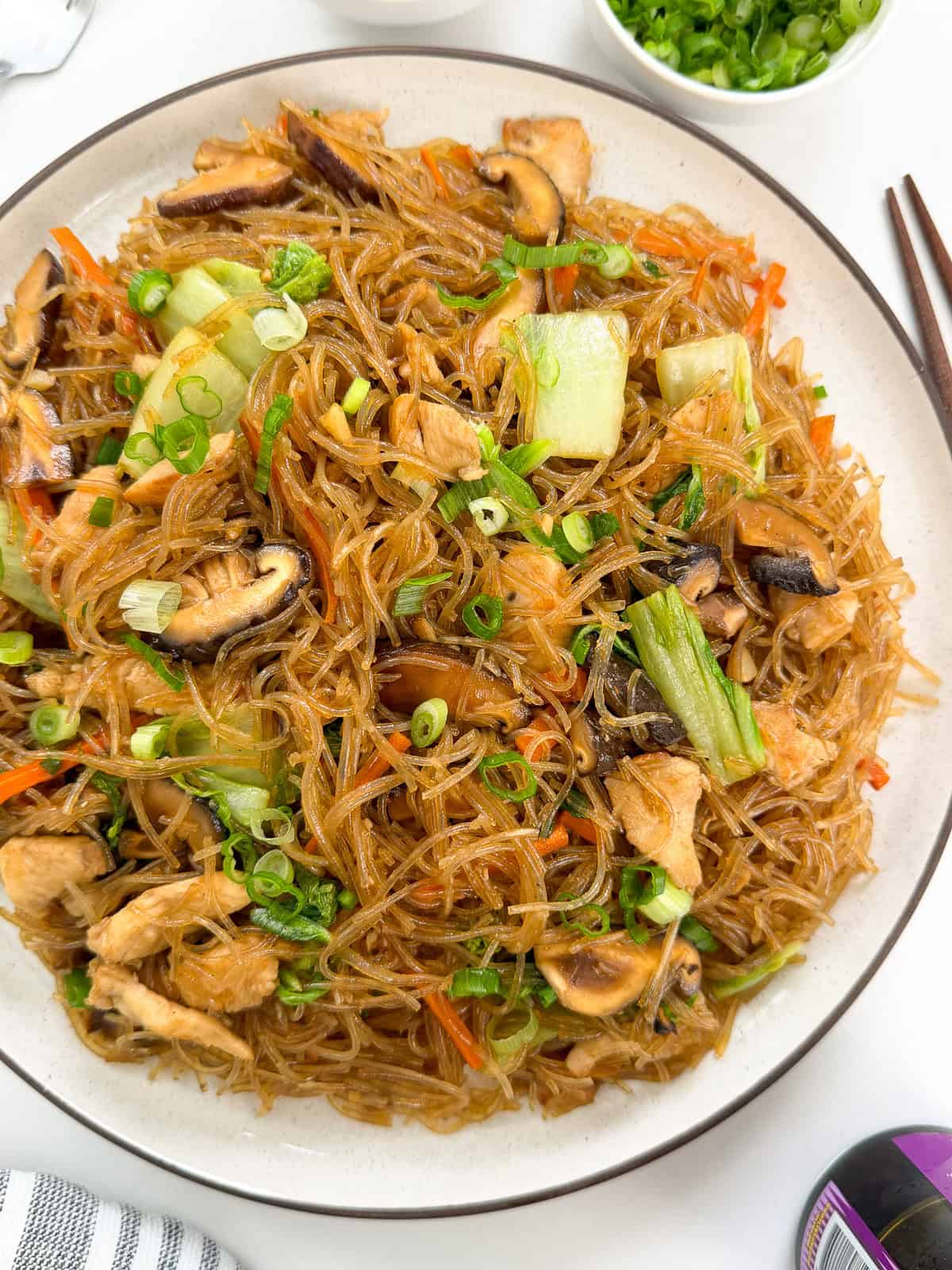 A plate of Stir-Fry Glass Vermicelli Noodles with sliced mushrooms, bok choy, carrots, chicken, and chopped green onions, arranged neatly and served with chopsticks on the side.