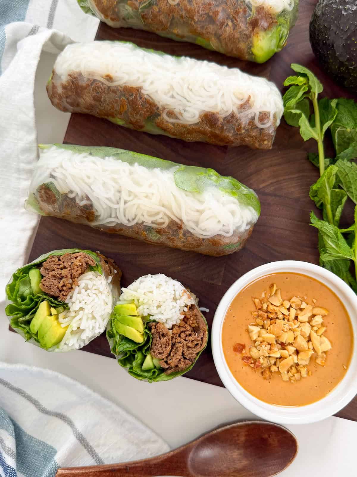 Three fresh Thai Beef Rice Paper Rolls and a halved roll are displayed on a wooden board. Nearby are mint leaves, avocado, and a bowl of peanut lime sauce garnished with chopped peanuts. A wooden spoon and striped cloth are also visible.