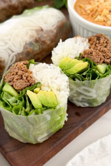 Thai Beef Rice Paper Rolls with Peanut Lime Sauce, cut in half and filled with rice, lettuce, avocado, and seasoned ground beef, are displayed on a wooden board with peanut lime dipping sauce topped with crushed peanuts.