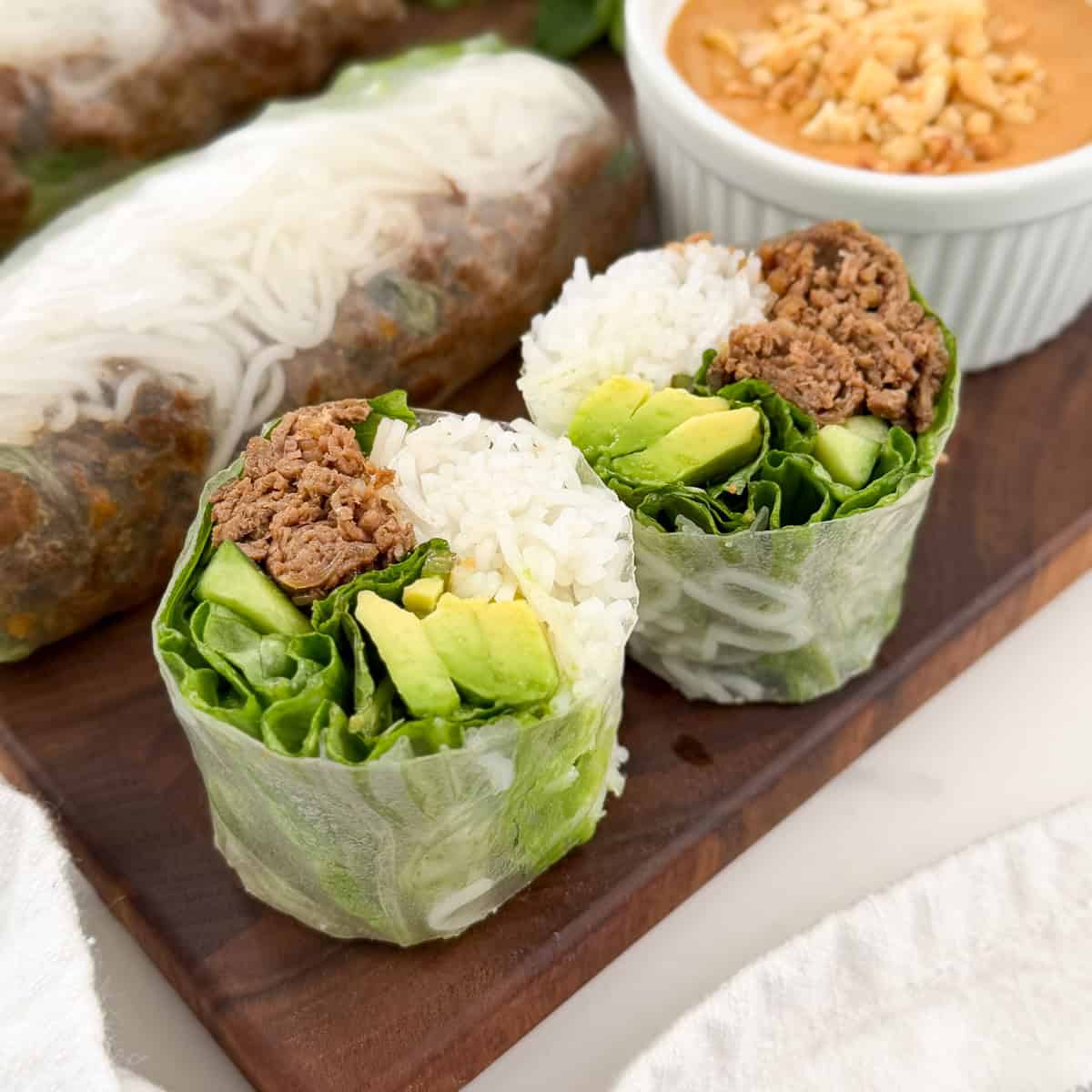 Thai Beef Rice Paper Rolls with Peanut Lime Sauce, cut in half and filled with rice, lettuce, avocado, and seasoned ground beef, are displayed on a wooden board with peanut lime dipping sauce topped with crushed peanuts.