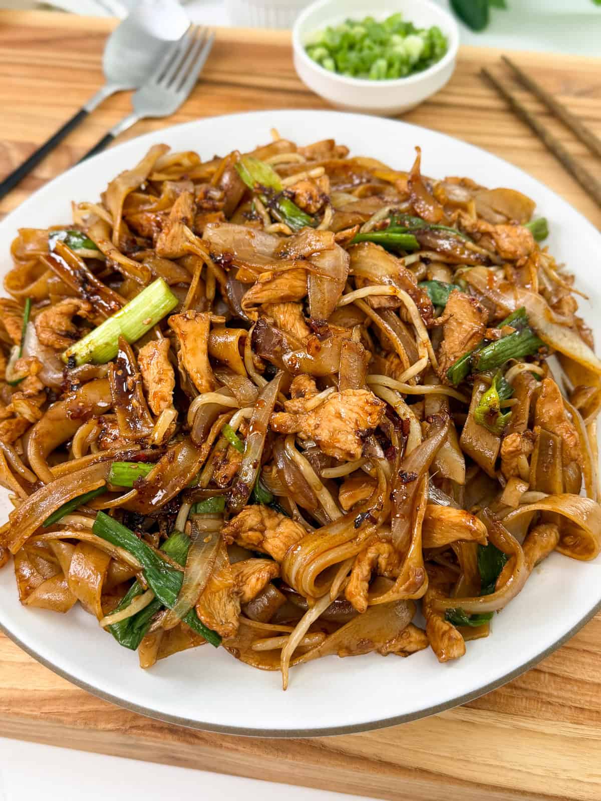A plate of Chicken Chow Fun featuring stir-fried flat rice noodles with chicken, green onions, bean sprouts, and a savory dark sauce is served on a white plate with chopsticks and a bowl of chopped green onions nearby.