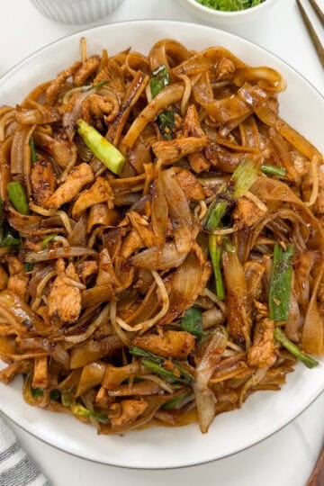 A plate of Chicken Chow Fun—stir-fried flat rice noodles with chicken, onions, green vegetables, and a dark savory sauce—is served with chopsticks, a spoon, and a napkin on a white table.