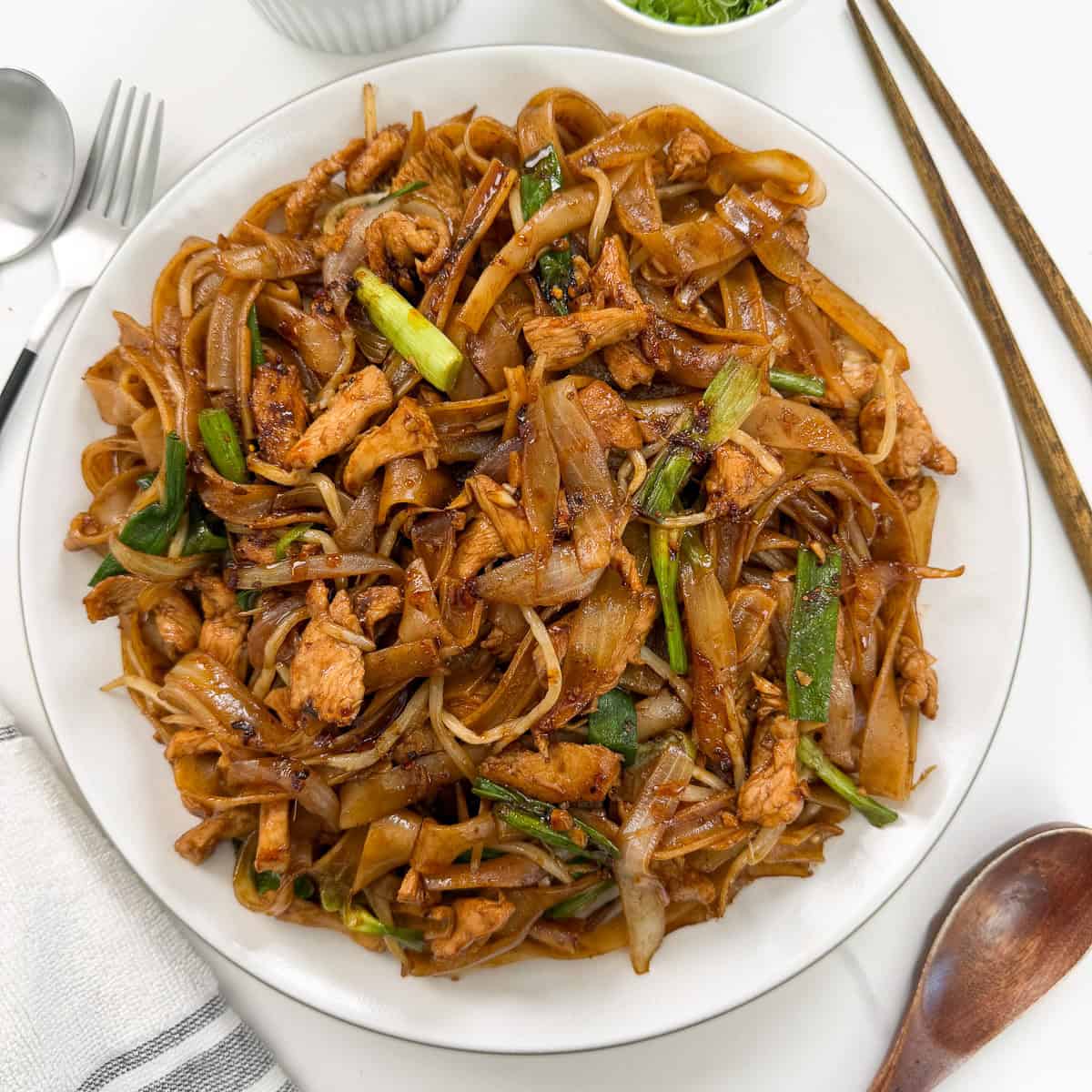 A plate of Chicken Chow Fun—stir-fried flat rice noodles with chicken, onions, green vegetables, and a dark savory sauce—is served with chopsticks, a spoon, and a napkin on a white table.