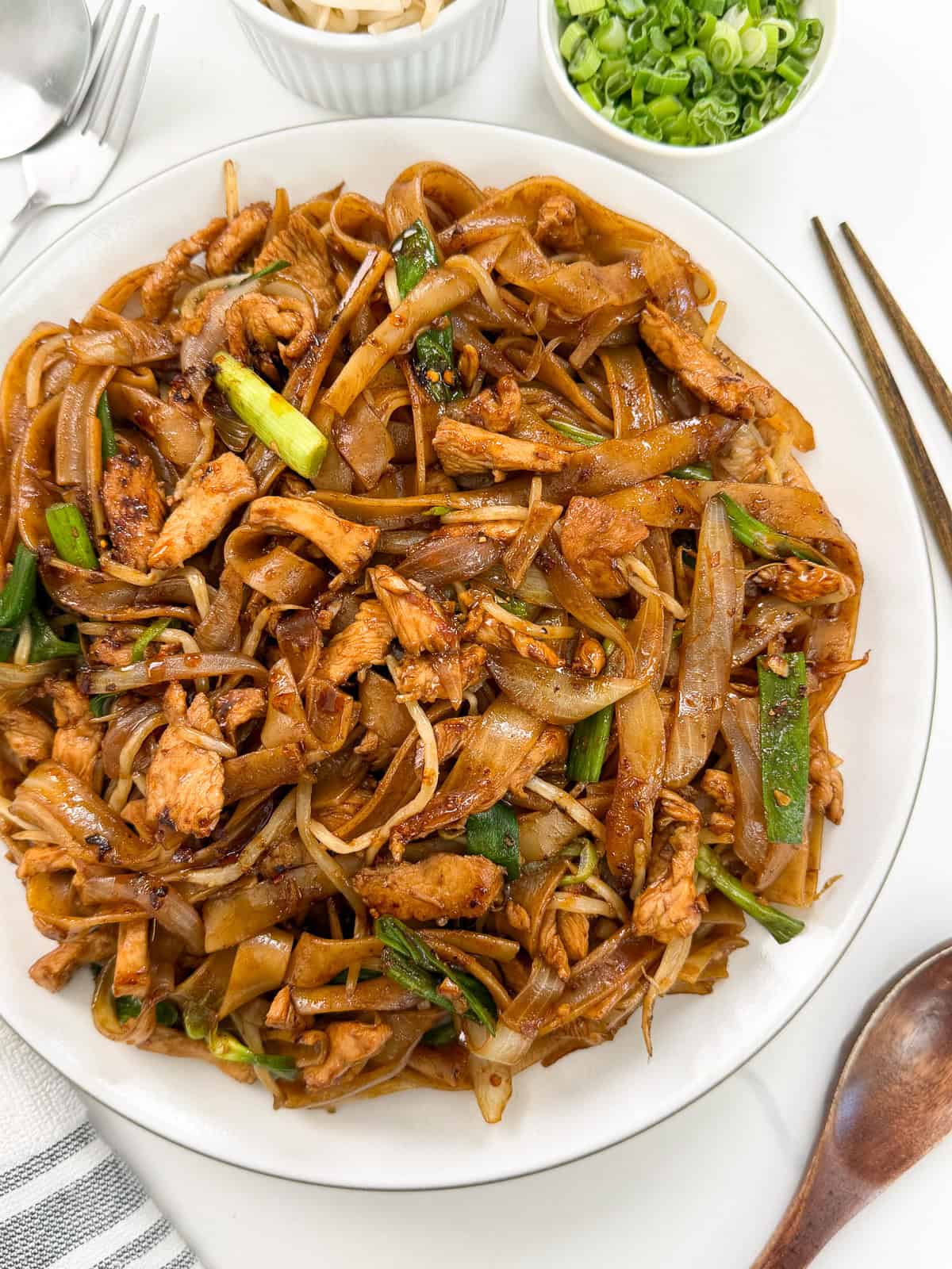 A plate of Chicken Chow Fun features stir-fried noodles with chicken, green onions, bean sprouts, and onions, garnished with chopped scallions. Small bowls of scallions and pickled vegetables are in the background.