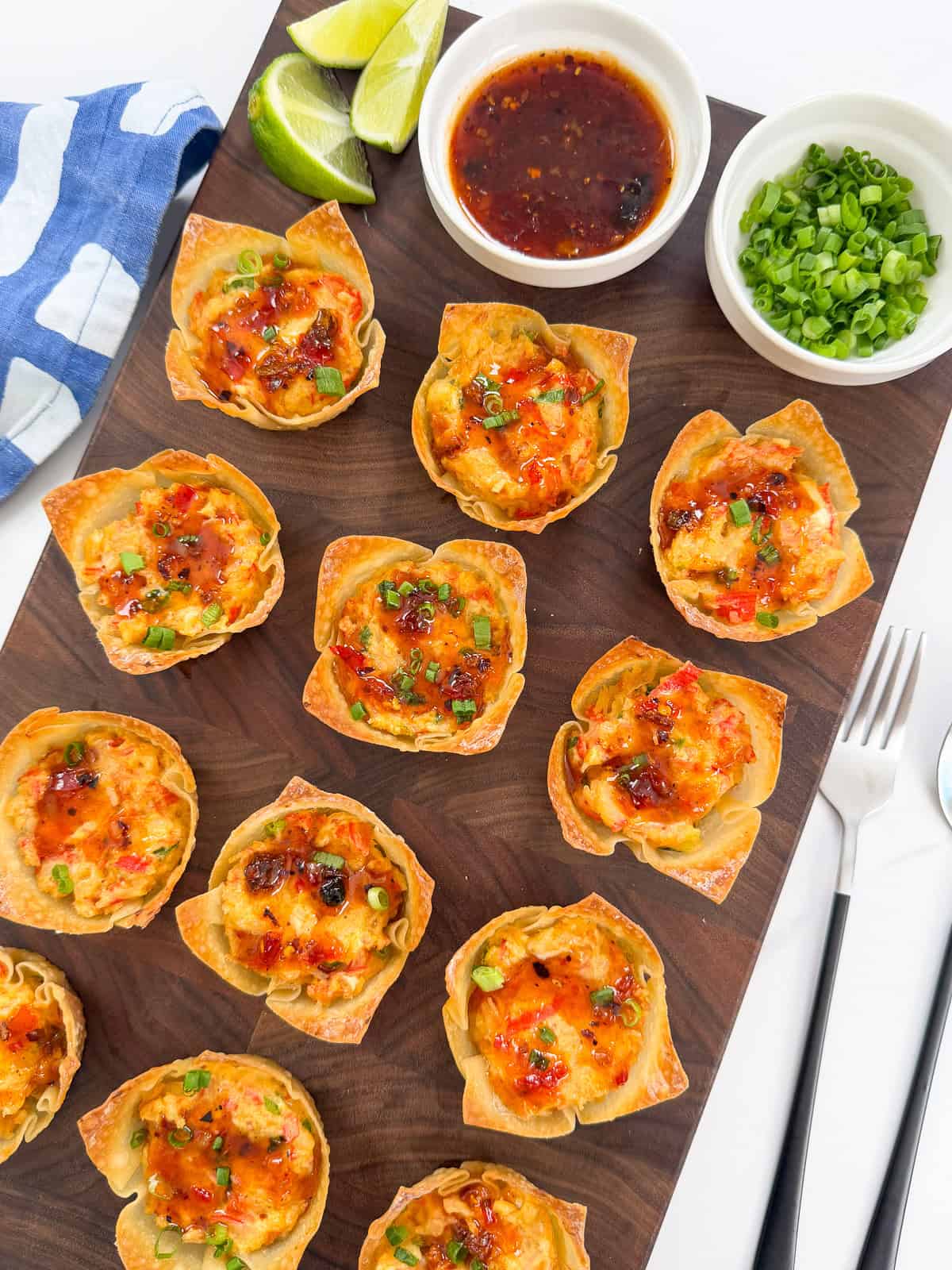 Mini Crab Rangoon Wonton Cups filled with a savory mixture, garnished with green onions, are arranged on a wooden board. Lime wedges, dipping sauce, and extra green onions are in small bowls nearby. A blue napkin and cutlery are on the side.
