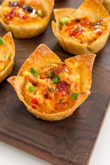 Crispy Crab Rangoon Wonton Cups filled with a cheesy, colorful mixture, garnished with chopped green onions and a drizzle of sauce, displayed on a wooden serving board.