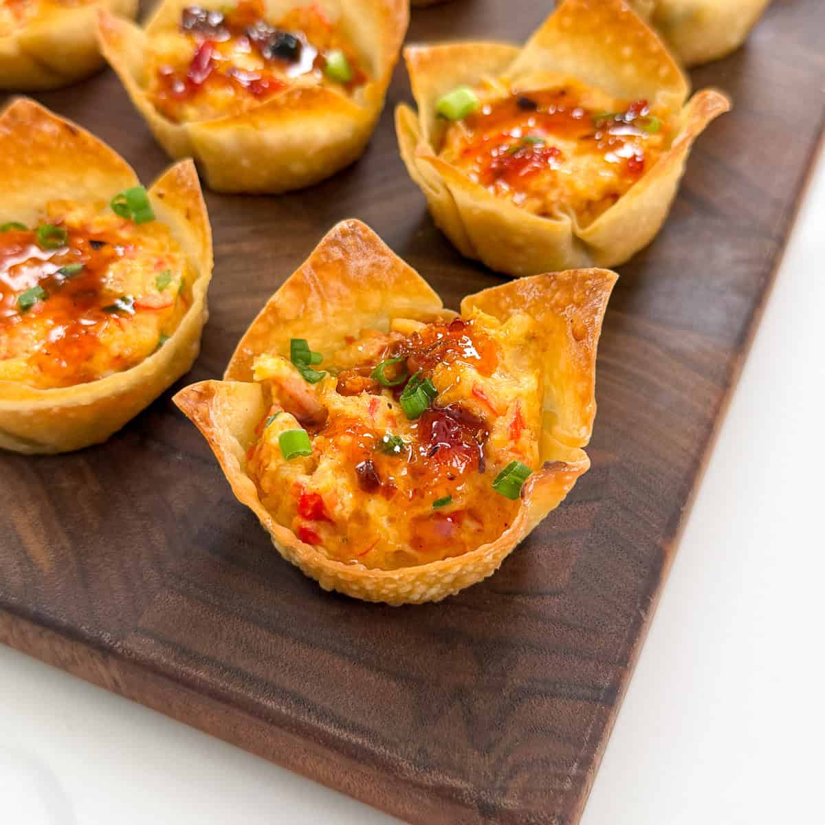 Crispy Crab Rangoon Wonton Cups filled with a cheesy, colorful mixture, garnished with chopped green onions and a drizzle of sauce, displayed on a wooden serving board.
