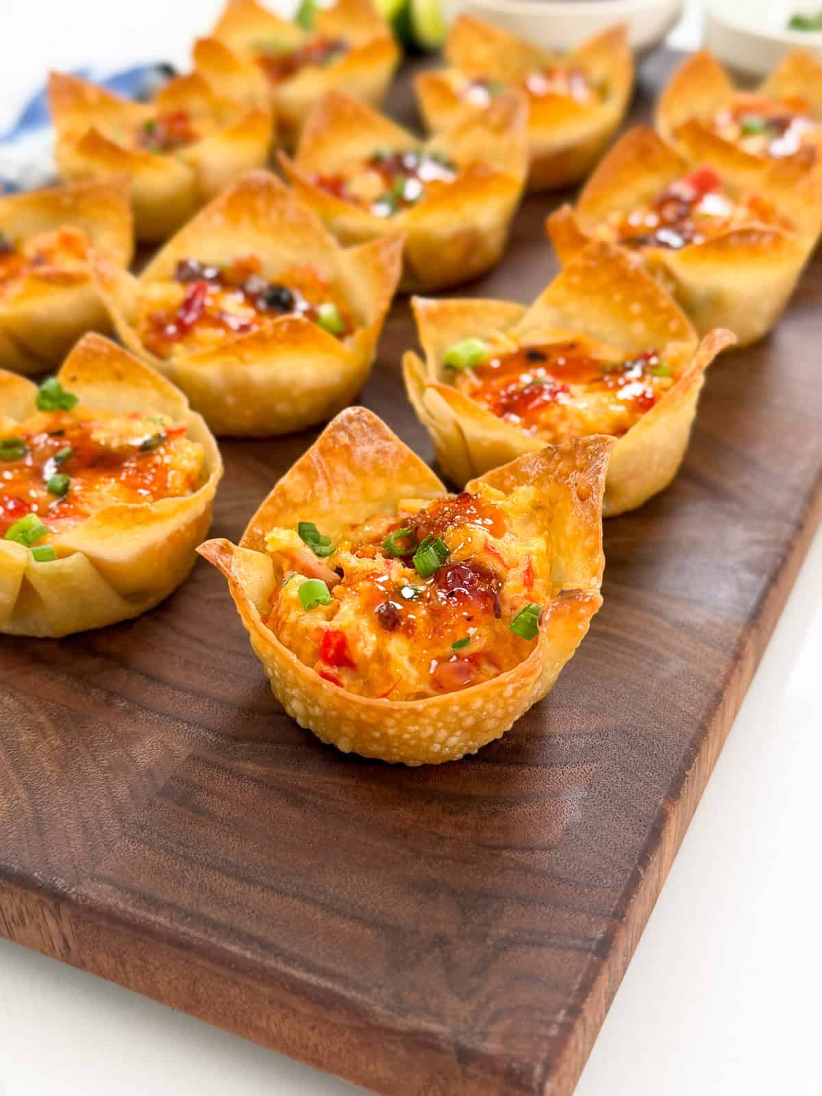 Crab Rangoon Wonton Cups: Mini wonton cups filled with a colorful mixture of vegetables, cheese, and savory crab, topped with sauce and chopped green onions, arranged neatly on a wooden serving board.