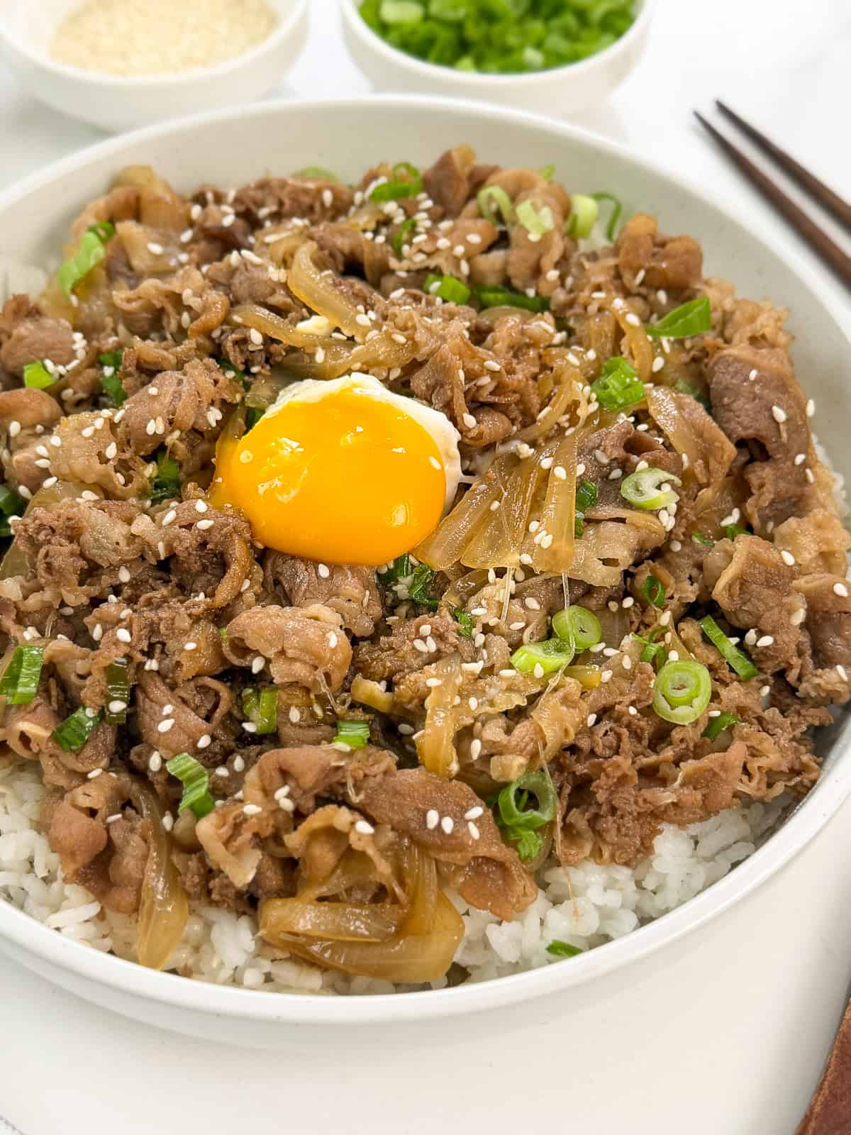 A bowl of Gyudon (Japanese Beef Rice Bowl) features sautéed beef, onions, chopped green onions, sesame seeds, and a raw egg yolk in the center atop steaming rice. Chopsticks and small dishes are in the background.