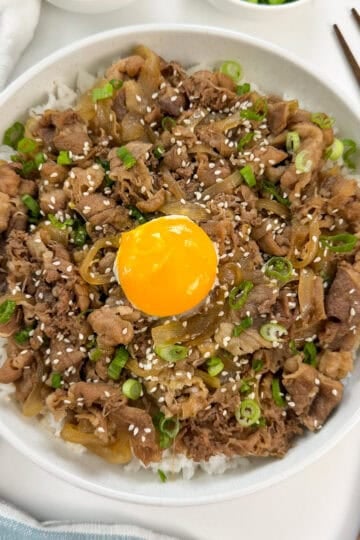 A bowl of Gyudon (Japanese Beef Rice Bowl) topped with cooked beef, caramelized onions, chopped green onions, sesame seeds, and a raw egg yolk in the center. Chopsticks and a spoon are placed beside the bowl.