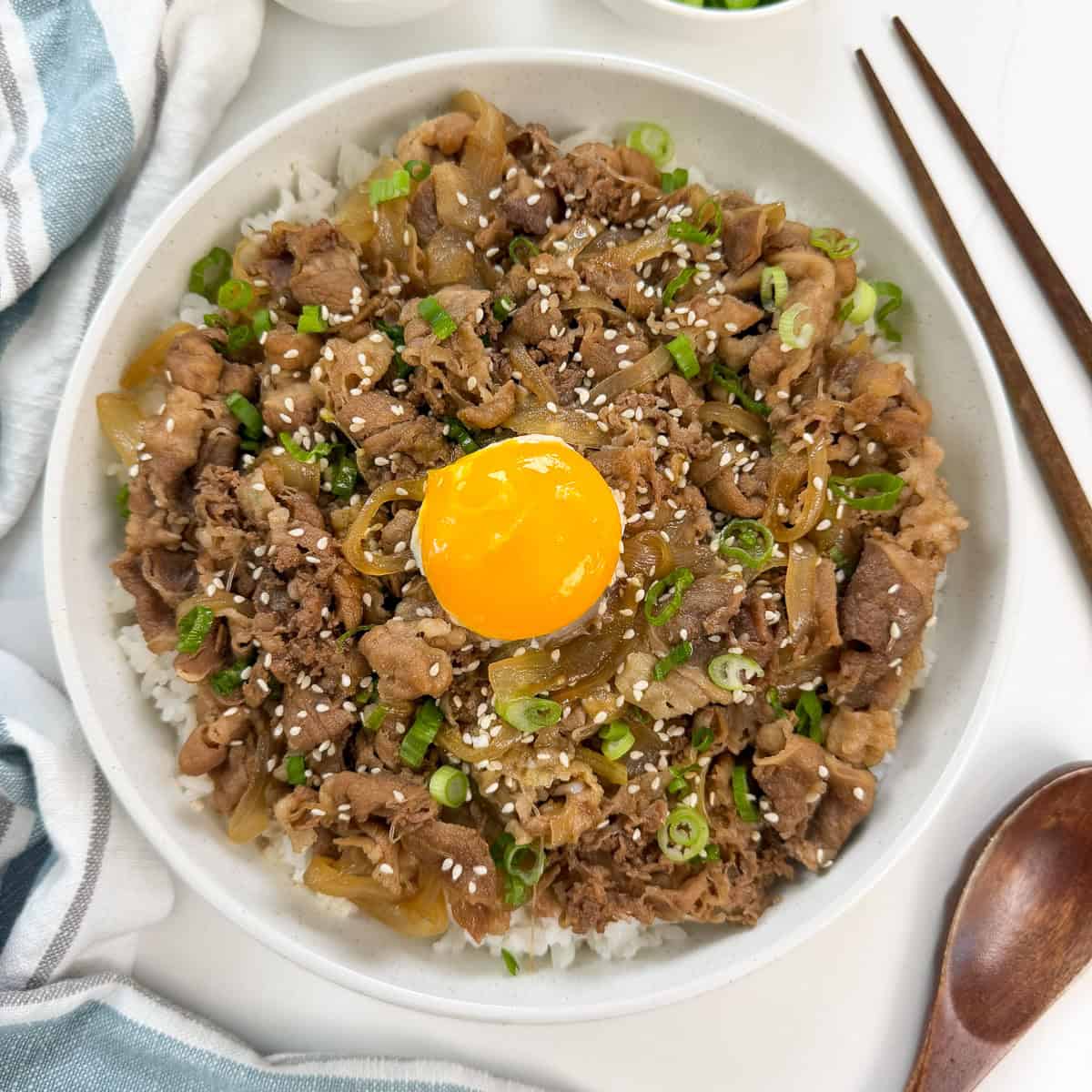 A bowl of Gyudon (Japanese Beef Rice Bowl) topped with cooked beef, caramelized onions, chopped green onions, sesame seeds, and a raw egg yolk in the center. Chopsticks and a spoon are placed beside the bowl.