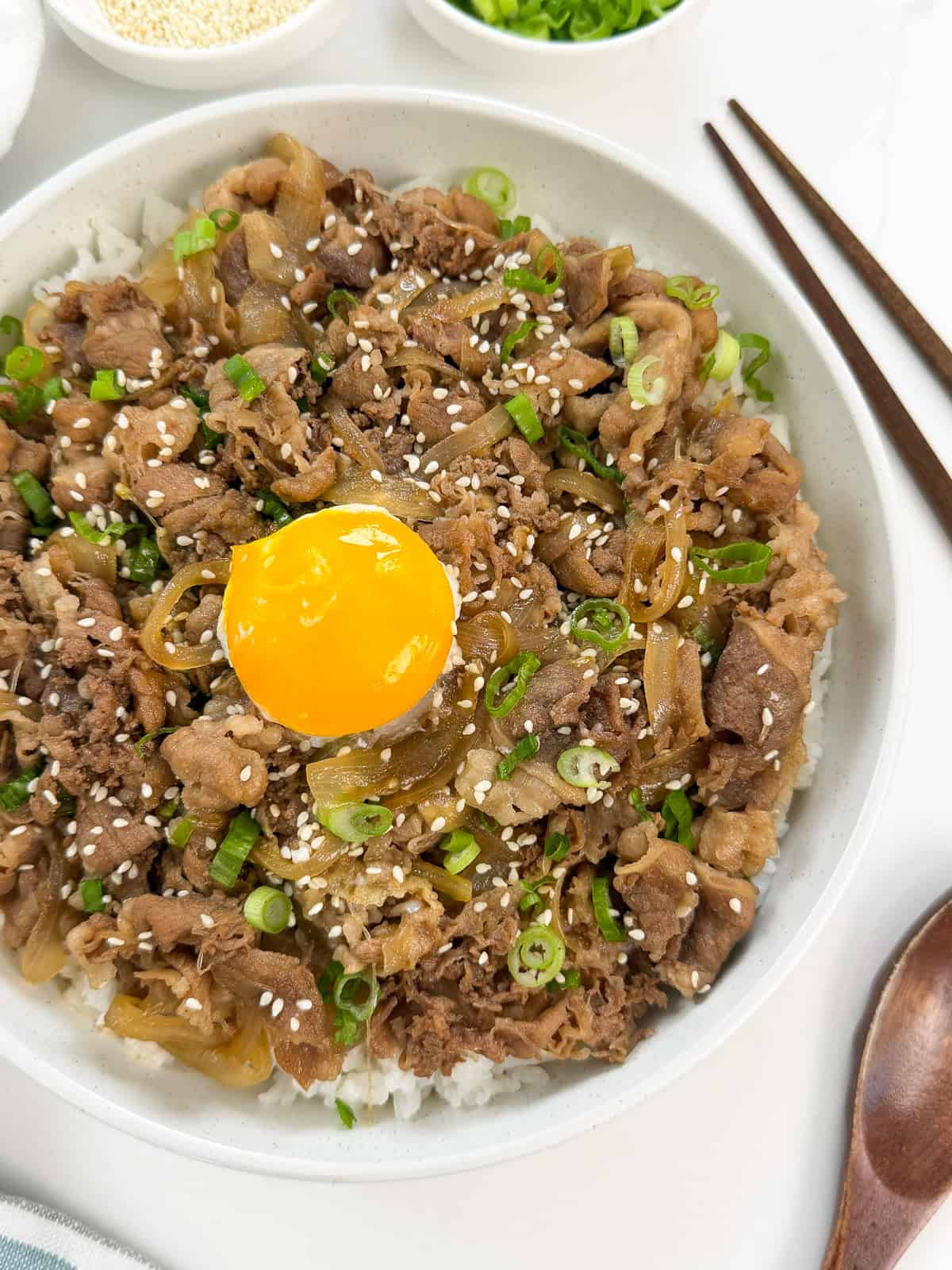 A bowl of rice topped with thinly sliced beef, onions, chopped green onions, sesame seeds, and a raw egg yolk in the center—this classic Gyudon (Japanese Beef Rice Bowl) is served with chopsticks and a spoon placed beside the bowl.