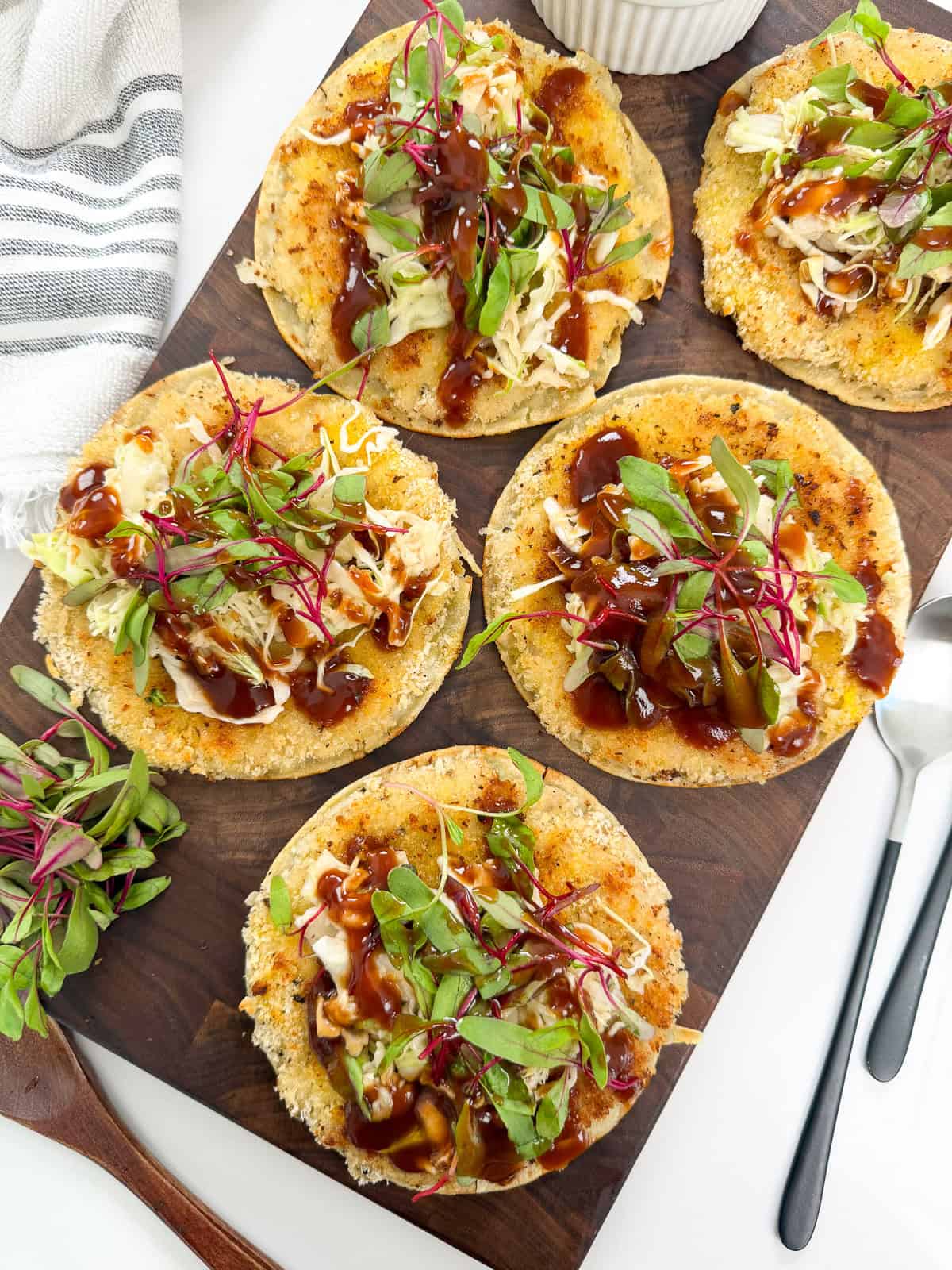 Five round tostadas, reminiscent of Katsu Chicken Smashed Tacos, are topped with shredded cabbage, leafy greens, and a dark sauce, artfully arranged on a wooden board with a small dish, forks, and a striped towel nearby.