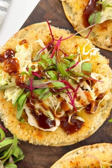 A close-up of a crispy tostada topped with shredded lettuce, microgreens, barbecue sauce, and cheese—reminiscent of flavors found in Katsu Chicken Smashed Tacos—served on a wooden platter.