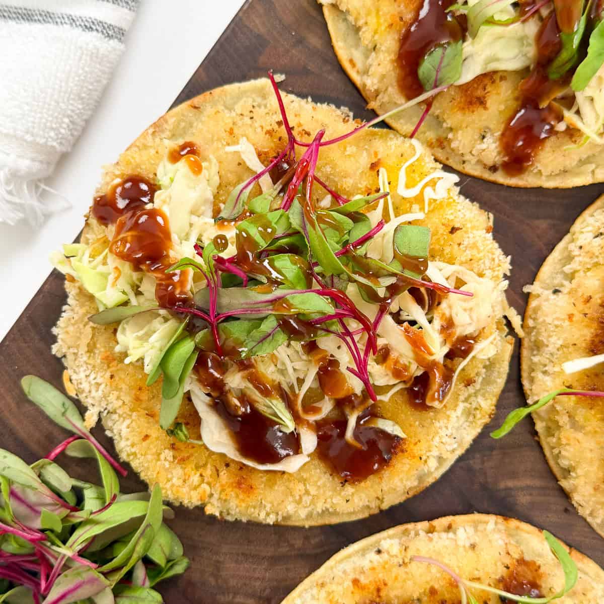 A close-up of a crispy tostada topped with shredded lettuce, microgreens, barbecue sauce, and cheese—reminiscent of flavors found in Katsu Chicken Smashed Tacos—served on a wooden platter.