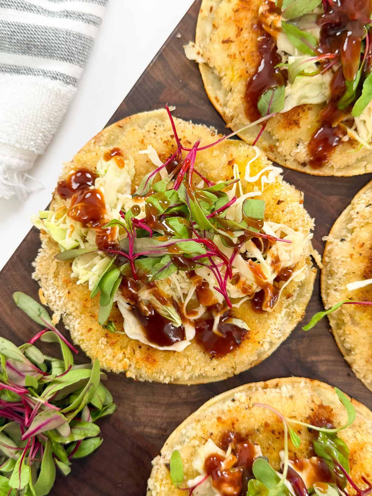 A close-up of crispy breaded tortillas filled with Katsu Chicken Smashed Tacos, topped with shredded lettuce, microgreens, and drizzles of dark sauce, arranged on a wooden board. A small pile of microgreens and a striped towel are nearby.