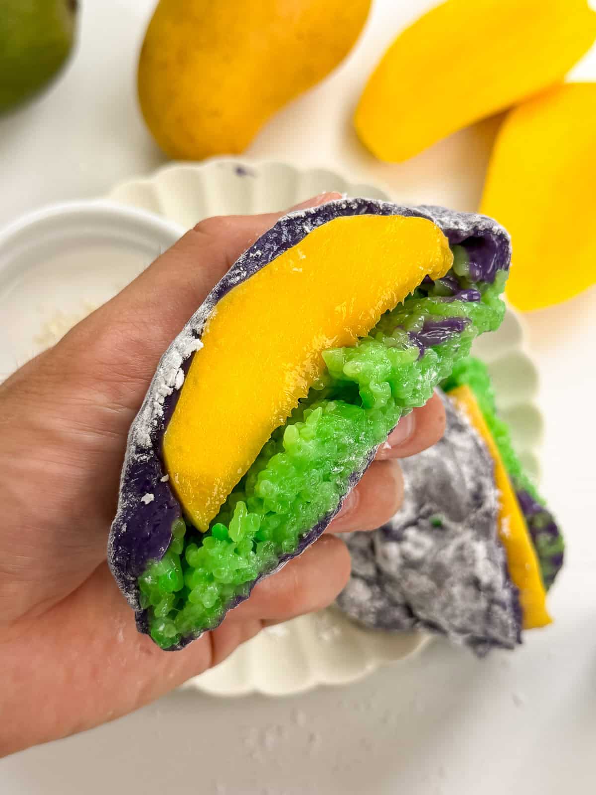 A hand holds a vibrant dessert featuring purple and white ube mochi, green sticky rice, and a slice of yellow mango. Sliced mangoes and another serving of mango sticky rice are on a white plate in the background.