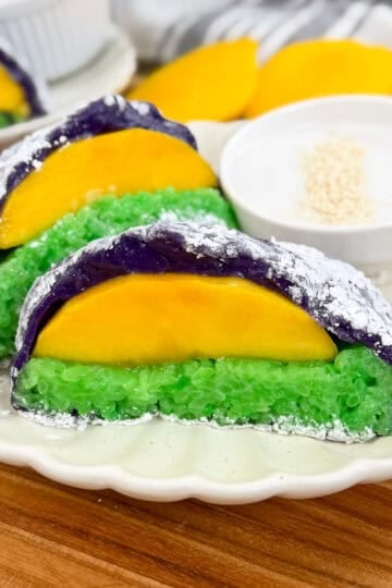 Three slices of colorful sticky rice cake with green and purple layers, inspired by Mango Sticky Rice Ube Mochi, are each topped with a slice of yellow mango. They are served on a plate with a small bowl of white dipping sauce and sprinkled sesame seeds.