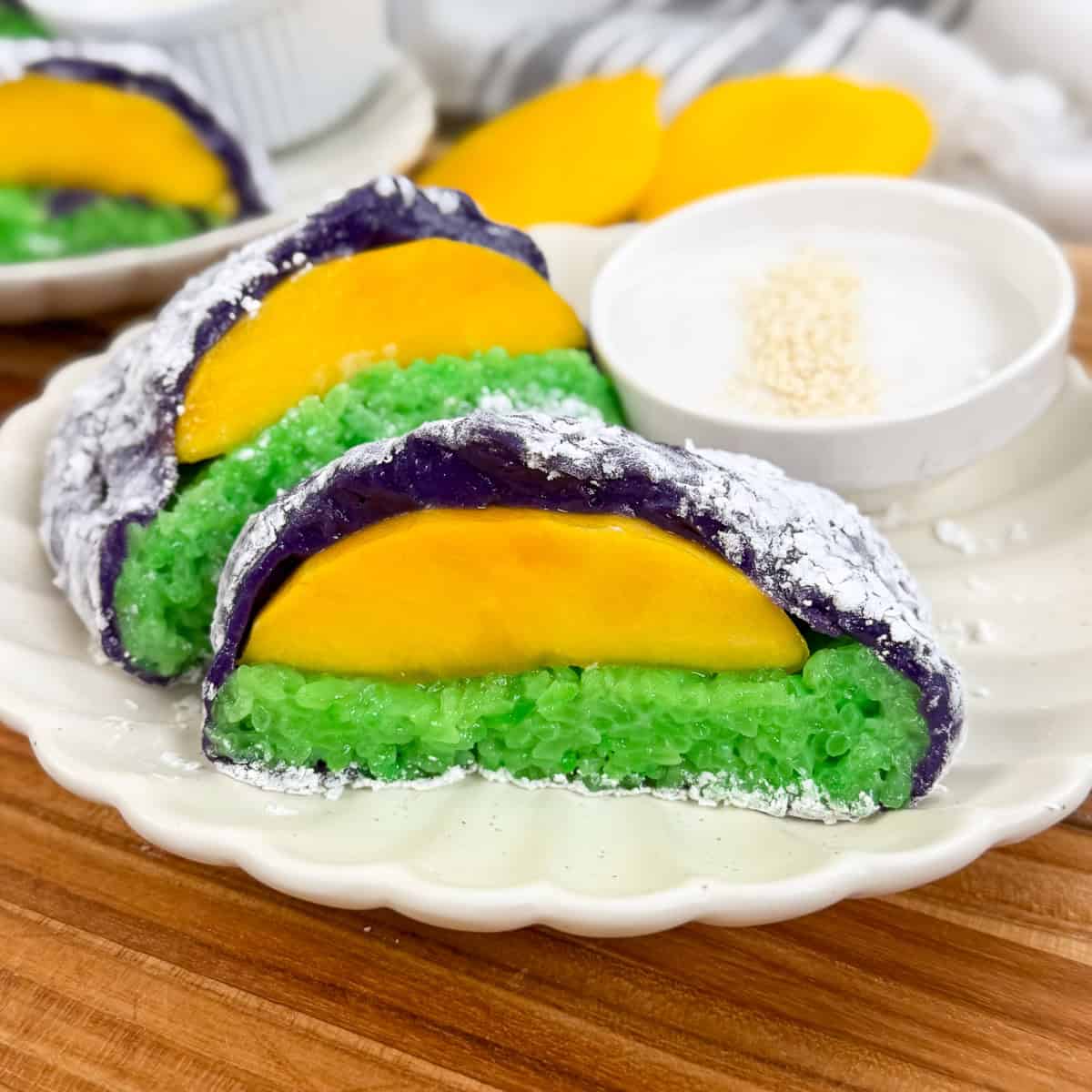 Three slices of colorful sticky rice cake with green and purple layers, inspired by Mango Sticky Rice Ube Mochi, are each topped with a slice of yellow mango. They are served on a plate with a small bowl of white dipping sauce and sprinkled sesame seeds.